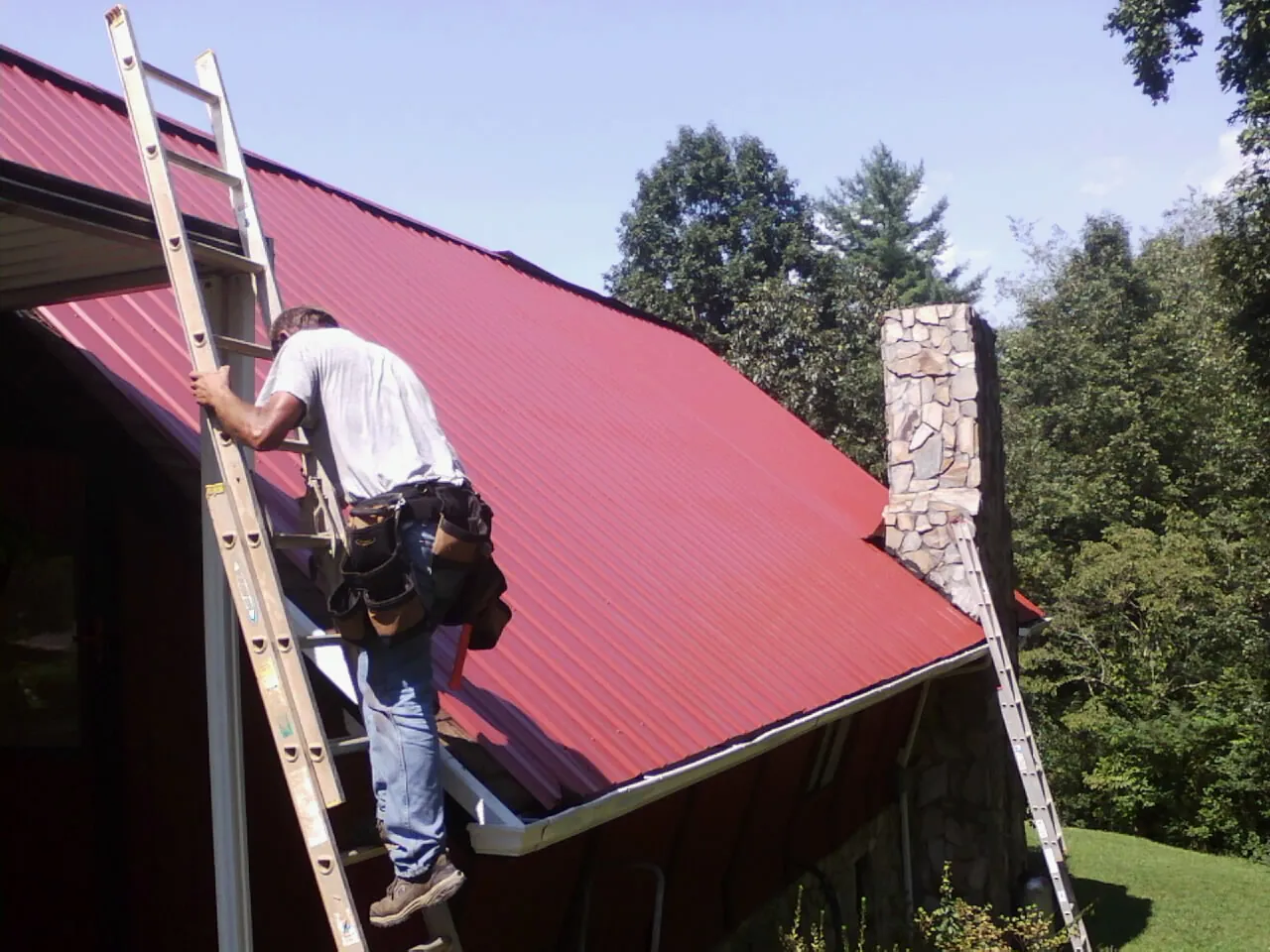 Roofer on a ladder beside a red metal roof and stone chimney — Soffit & Fascia Repair by Roofing Brothers in Gleneden Beach